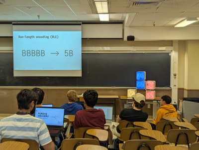 projector slide with heading 'run-length encoding (RLE)' and large text 'BBBBB -> 5B'. people facing projector slide.