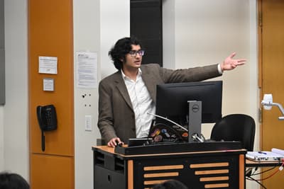 man wearing white shirt and brown coat at a lectern points to the right